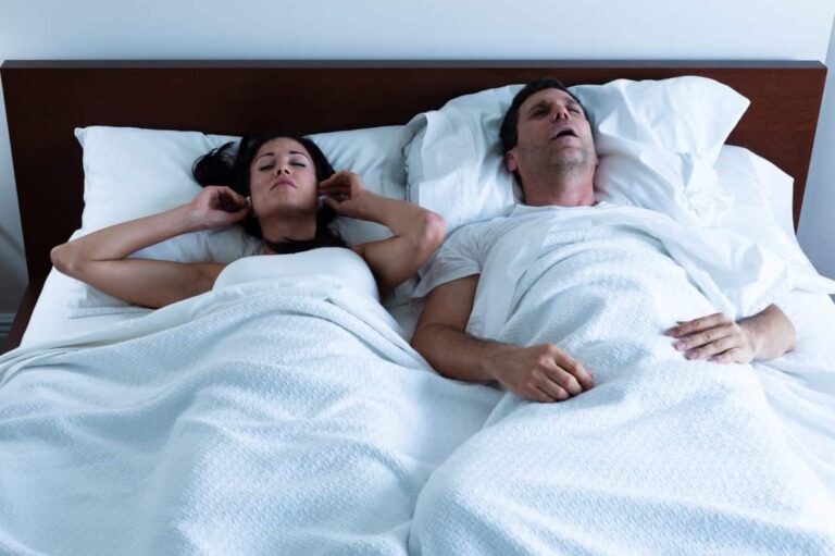 snoring-solutions A heterosexual couple in bed. The man on the right has his mouth open as though snoring. The woman on the left is placing ear plugs into her ears.