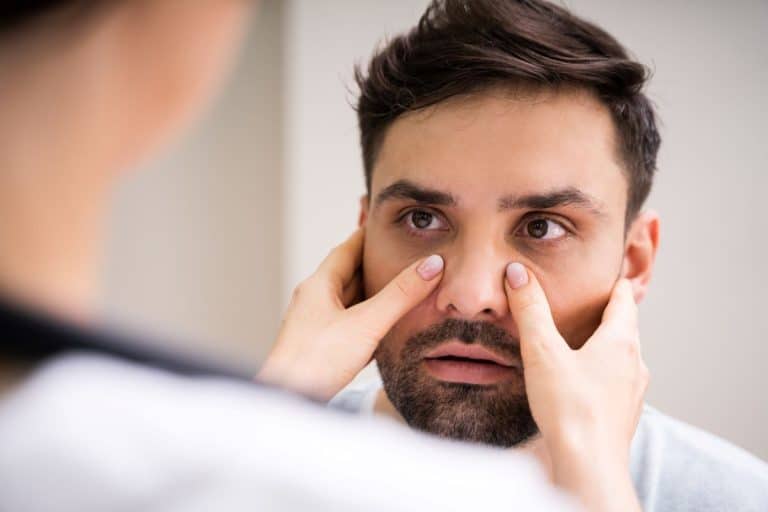 Rhinitis Sufferer Blurry back shoulder and face of a person pressing their thumbs on either side of the bridge of the nose of a man who is looking at them.